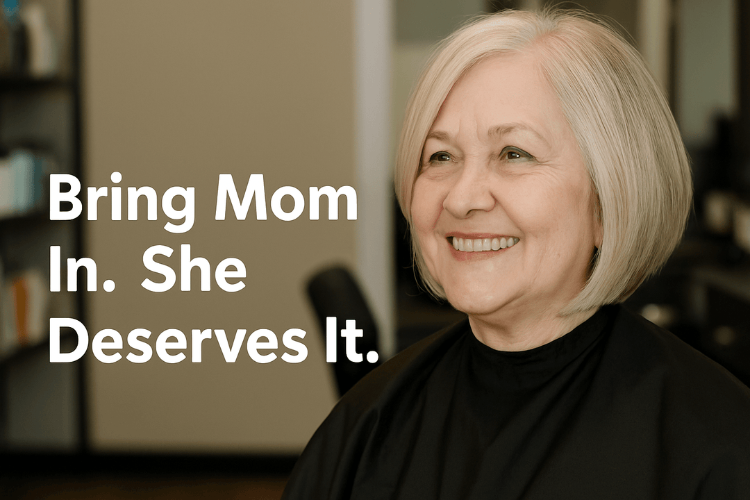 Smiling older woman with a fresh short bob haircut sitting in a salon chair, with bold text on the left reading ‘Bring Mom In. She Deserves It.’ inside The Warehouse Salon.